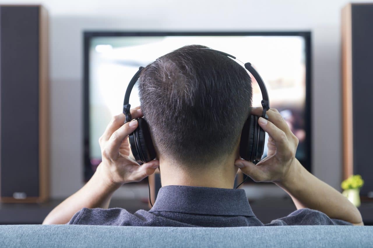 Back side of an man wearing black heaphones in front of blurry out-of-focus television and home entertainment system background