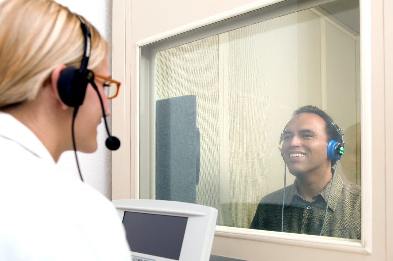 Man getting a hearing test, sitting in a booth, while a doctor administers the test outside of the booth