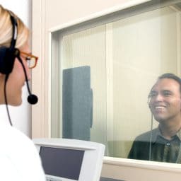 Man getting a hearing test, sitting in a booth, while a doctor administers the test outside of the booth